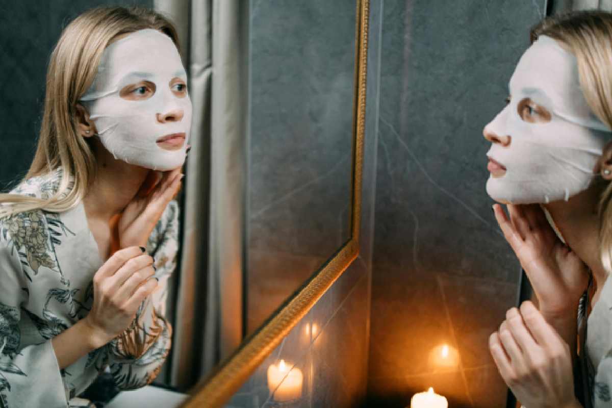 Woman wearing a white sheet face mask looking in bathroom mirror with candles during nighttime skincare routine.